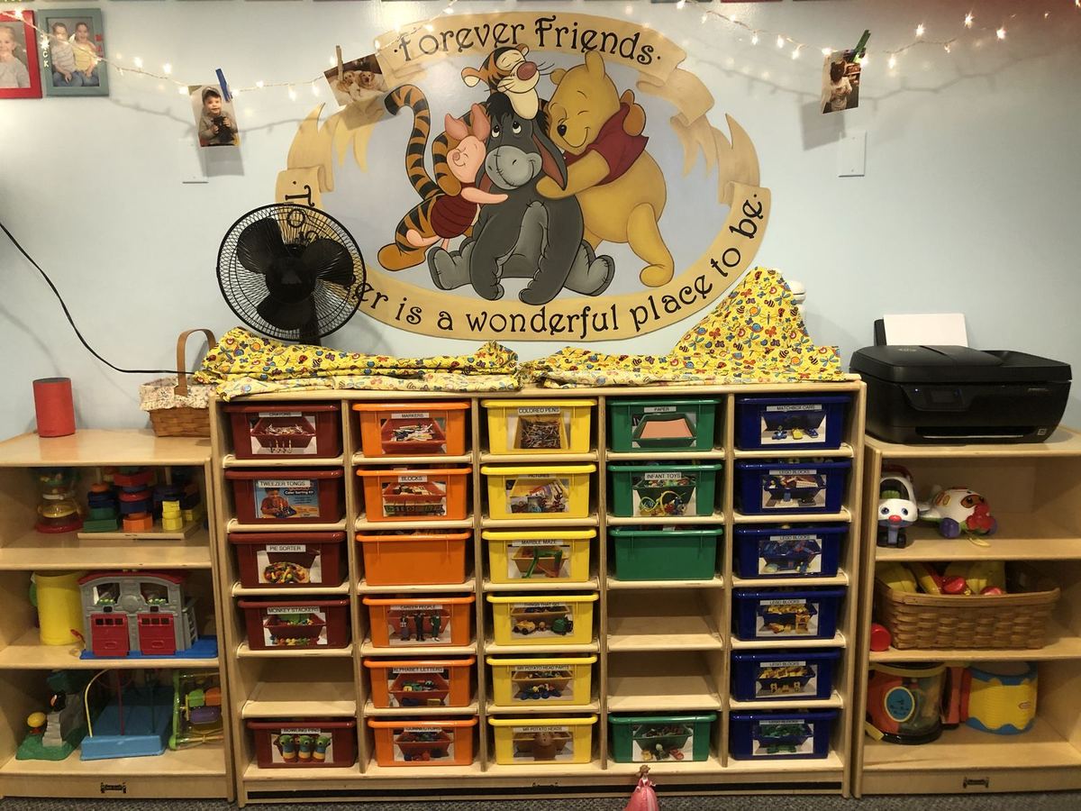 The main activity room: a wall of colored toy bins under a Winnie the Pooh mural that reads “Forever friends, together is a wonderful place to be,” with string lights and photos above